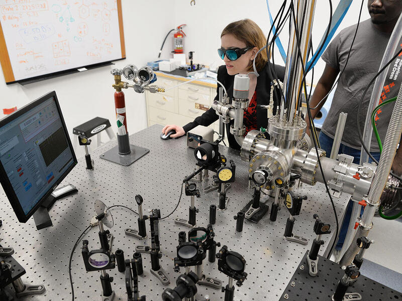 A woman with goggles works on a computer surrounded by lab equipment on a large table. A man in a gray shirt stands to her left.