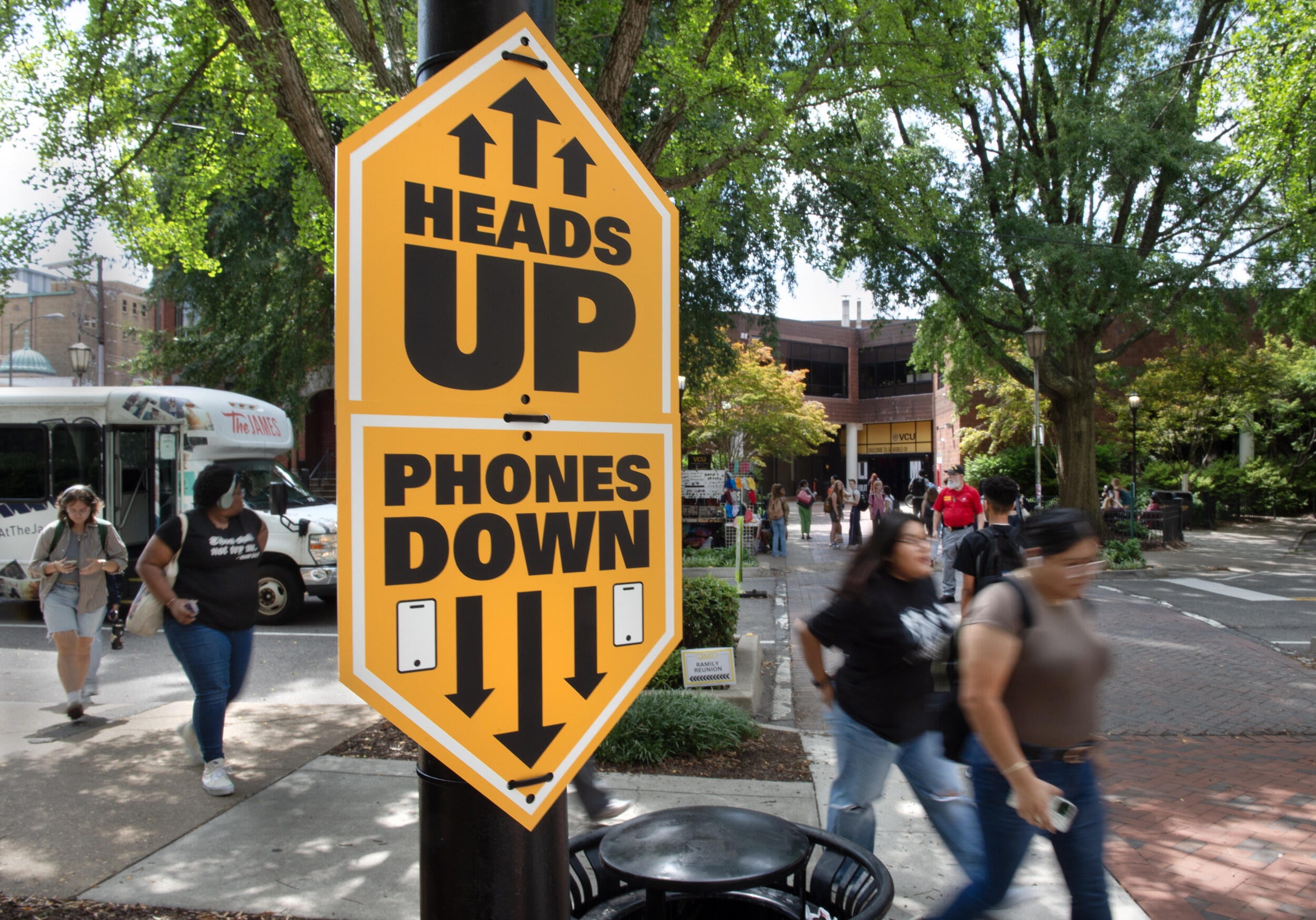 A photo of a sign by a cross walk that says \"HEADS UP\" \"PHONES DOWN\" with arrows pointing up and down. Behind the sign people are walking across the street and down a sidewalk. 