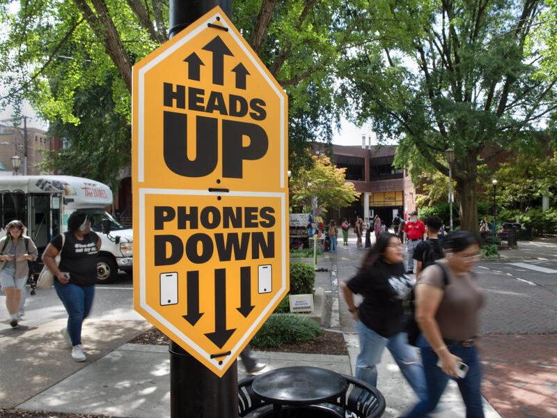 A photo of a sign by a cross walk that says \"HEADS UP\" \"PHONES DOWN\" with arrows pointing up and down. Behind the sign people are walking across the street and down a sidewalk. 