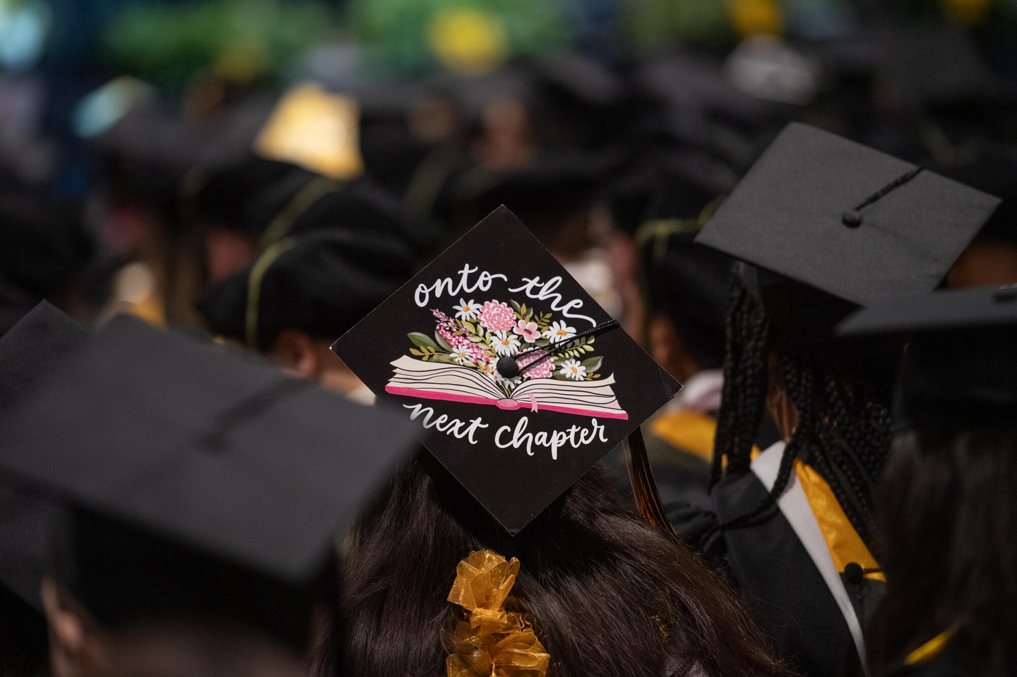A graduate wears a cap decorated with flowers and book that says \"Onto the next chapter.\"