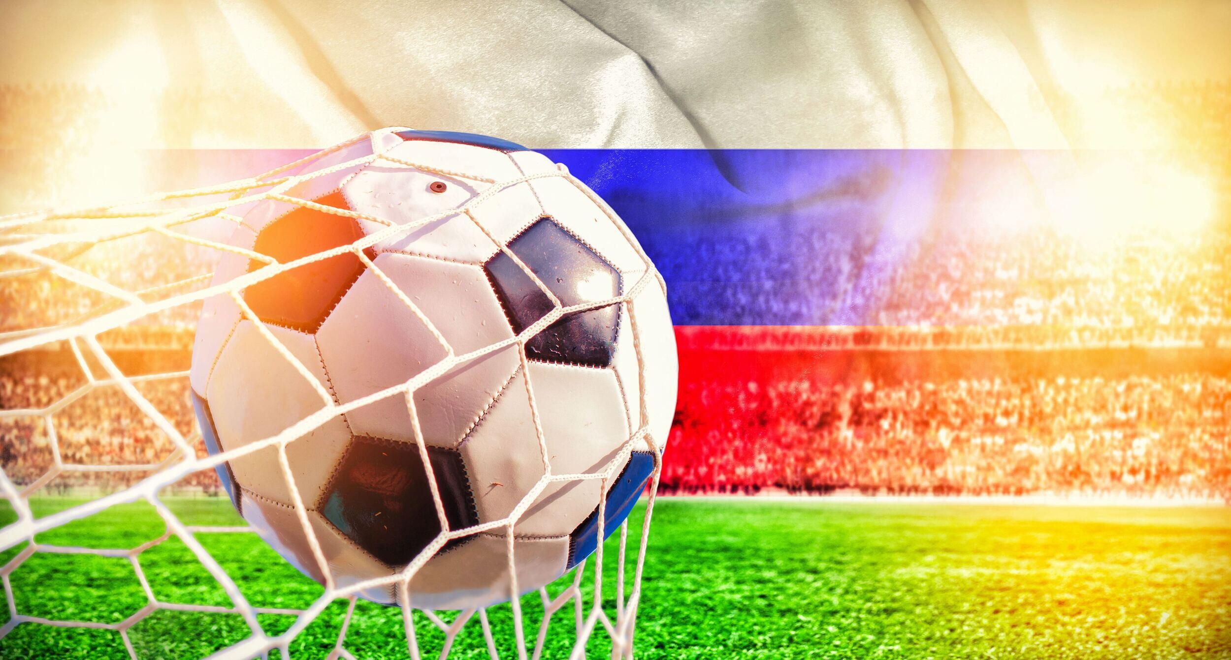 A soccer ball hitting the back of a net on a field with a Russian flag on top of the crowd of spectators 