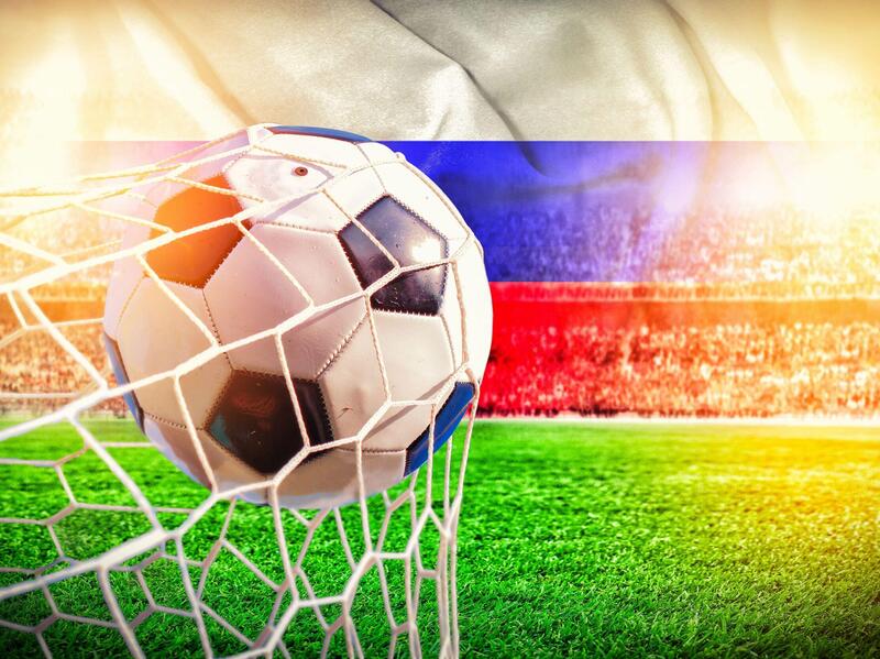 A soccer ball hitting the back of a net on a field with a Russian flag on top of the crowd of spectators 