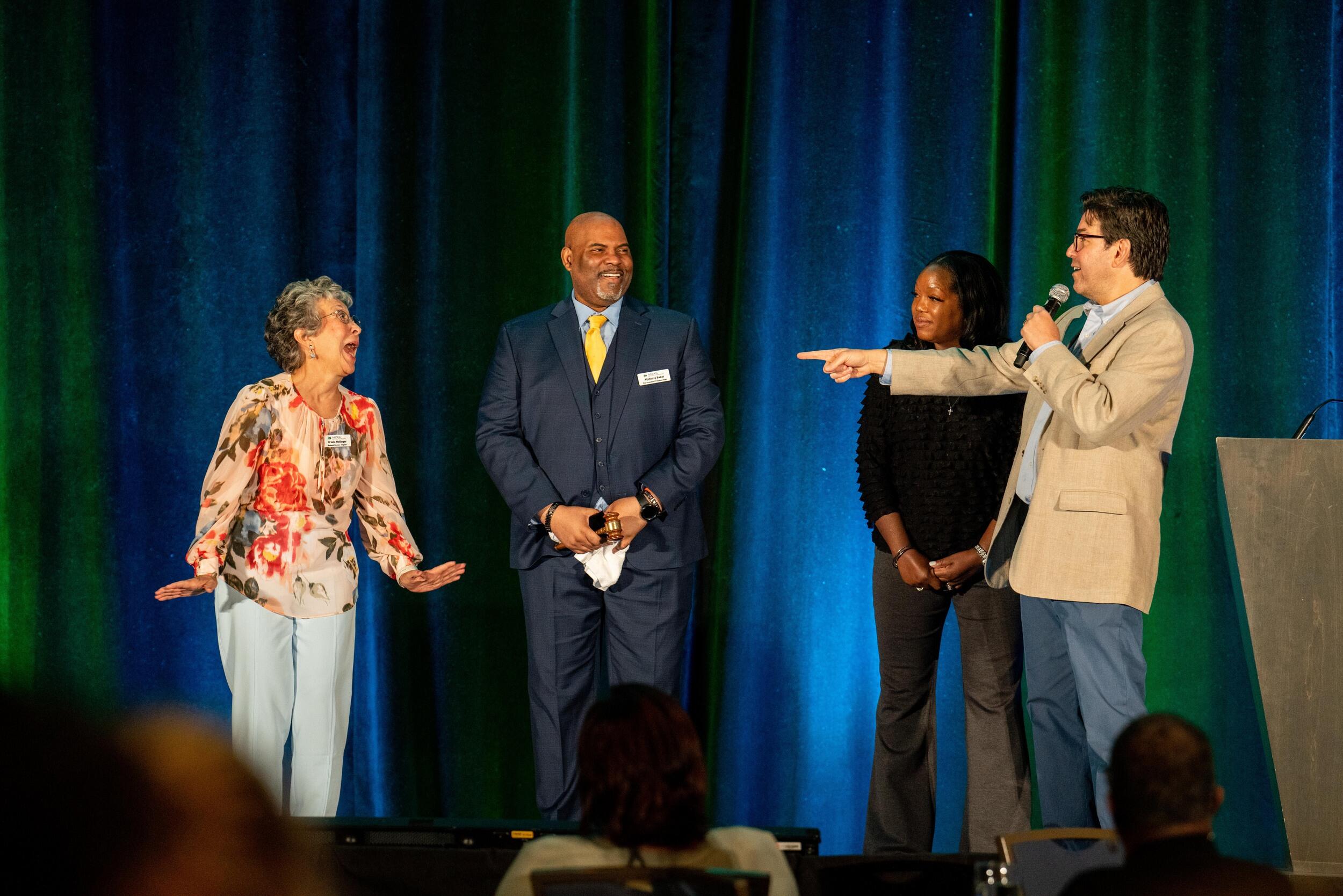 A photo of four people on stage. The person furthest to the right is pointing with his left arm the furthest person on the left. 