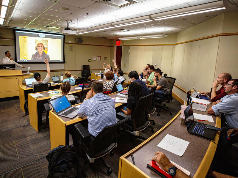 Students in a classroom interact with a teacher and a person participating through video conferencing.