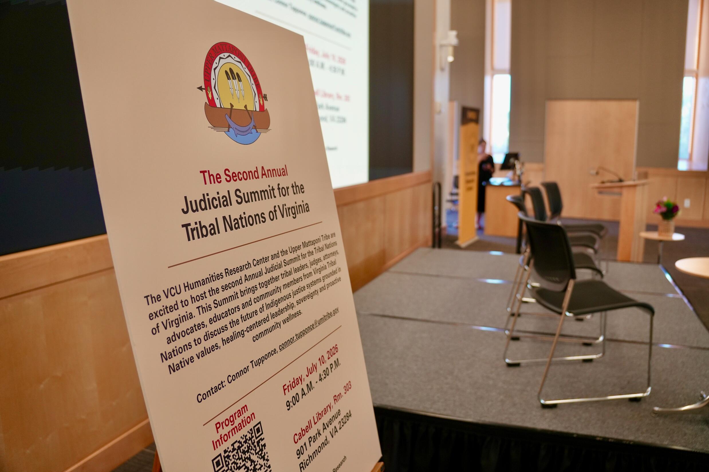 A photo of a sign next to a stage that says \"The Second Annual Judicial Summit for the Tribal Nations of Virginia.\"