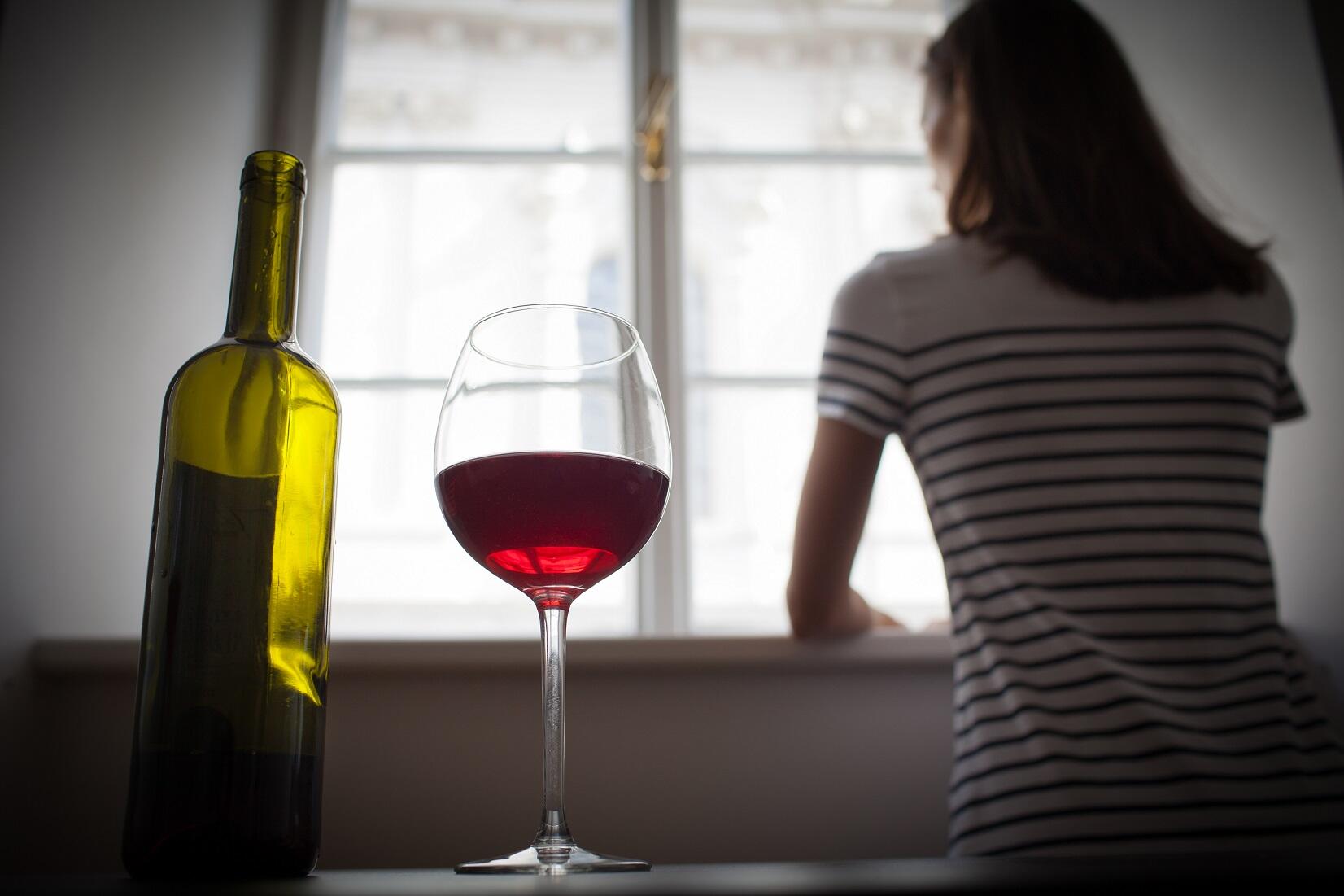 A green bottle and a glass half-full of red wine sit in the foreground. In the background, a woman is looking out a window.