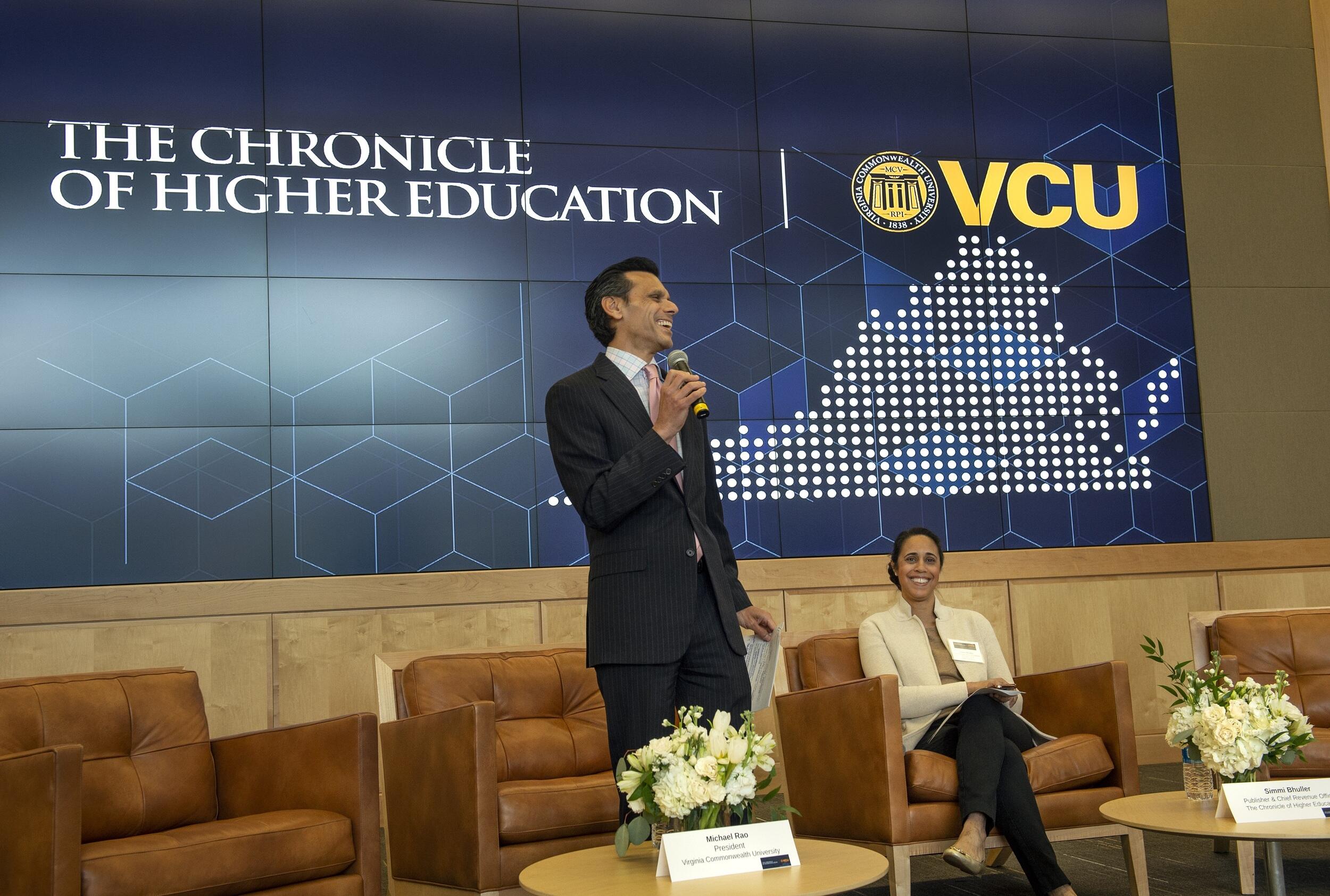 A man standing with a microphone in front of a screen that says \"THE CHRONICLE OF HIGHER EDUCATION\" next to an illustration of Virginia with the letters \"VCU\" above hit. Next to him a woman is sitting in a brown chair and smiling. 