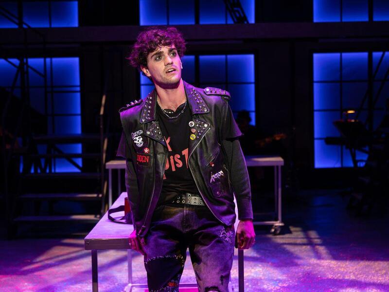 A photo of a man wearing a black leather jacket with studs and patches standing on a theater stage. 