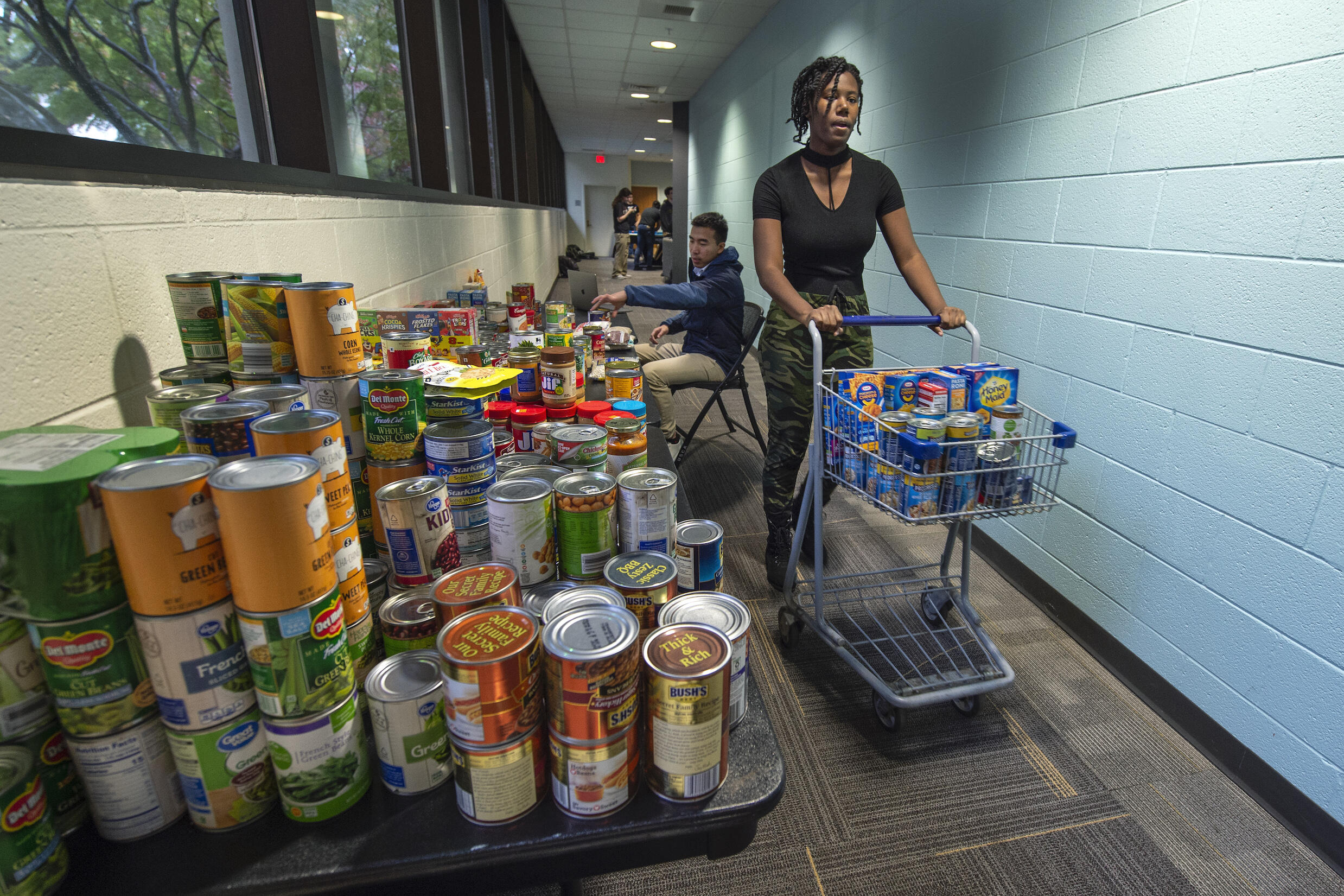A person pushes a grocery cart full of donated items. Cans and other donated food are stacked on a table.