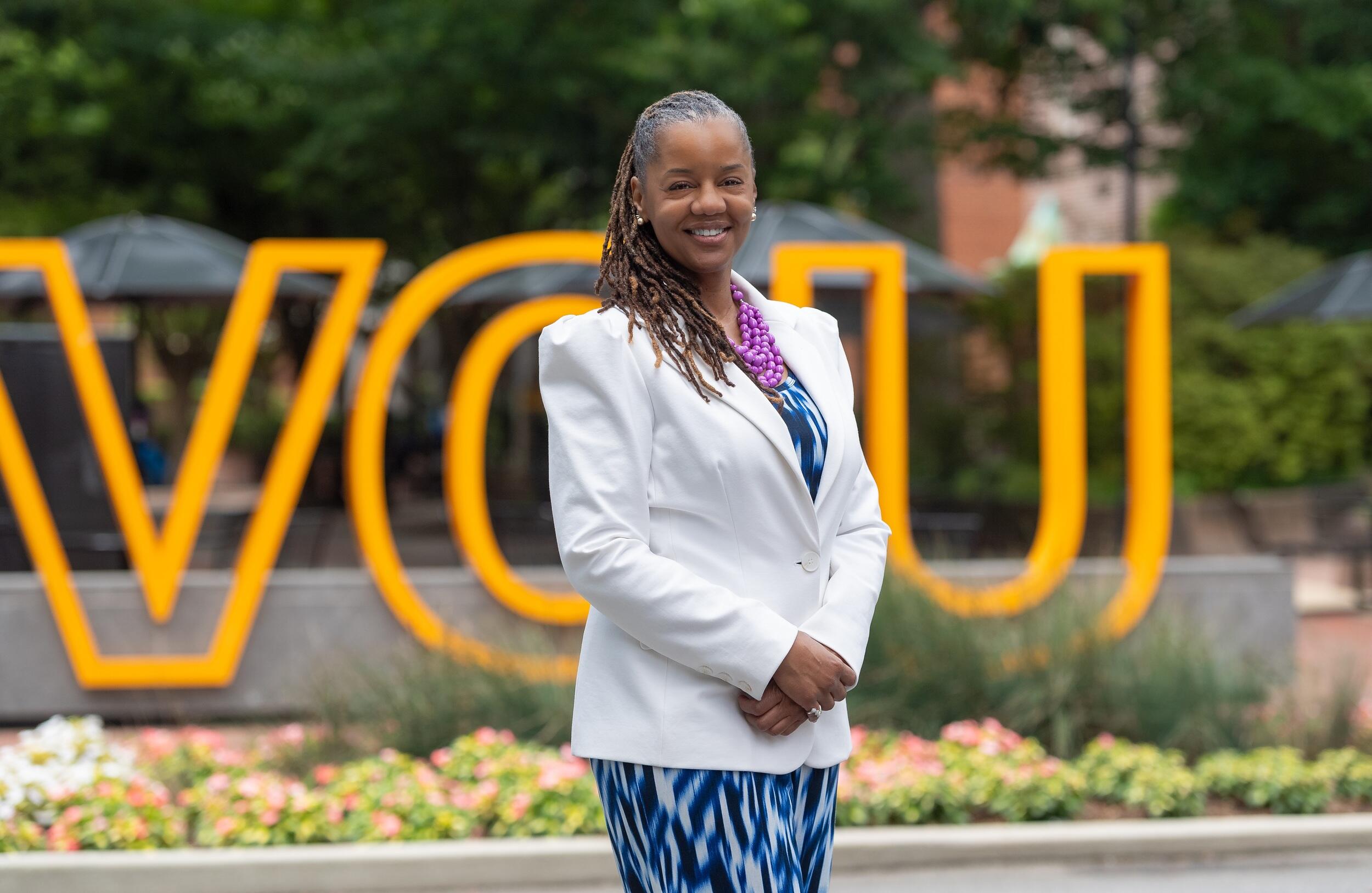 A photo of a woman from the thighs up standing in front of a sign that spells out \"VCU\" in giant yellow letters. 