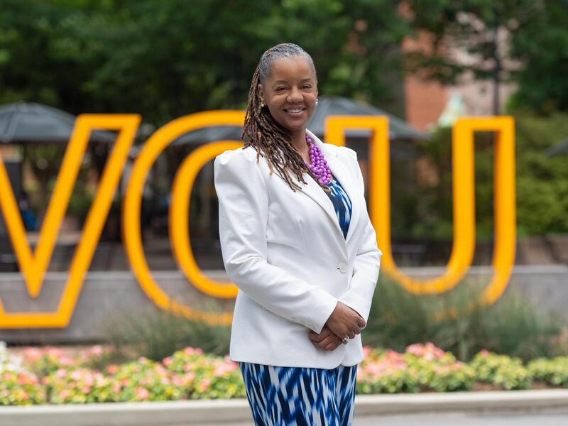A photo of a woman from the thighs up standing in front of a sign that spells out \"VCU\" in giant yellow letters. 