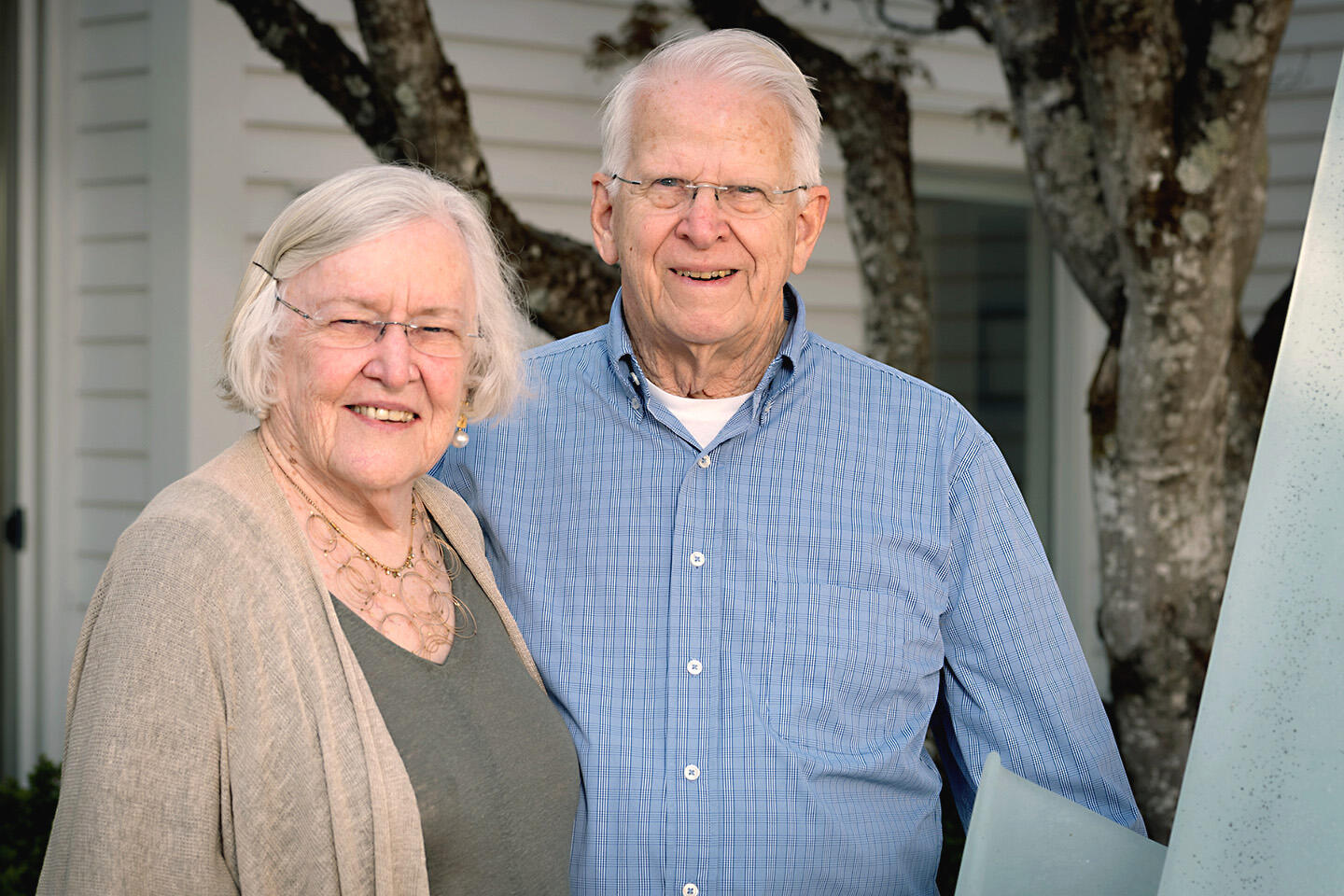 A photo of a woman and a man from the waist up. They are standing outside in front of a tree. Behind the tree is a house. 