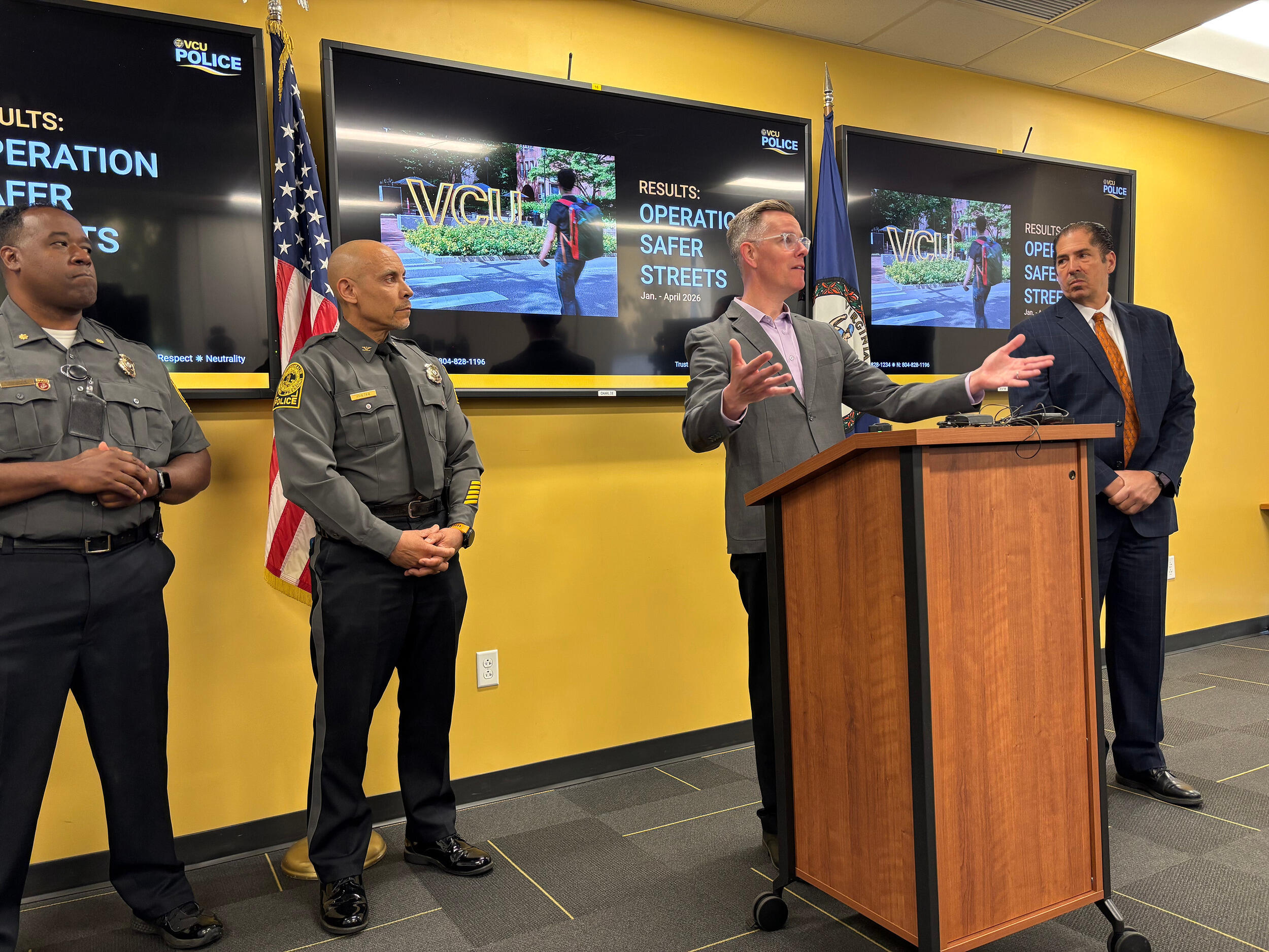 A photo of a mand standing behind a podium. On his right is a man in a dark blue suit. On the man's left are two police officers. 