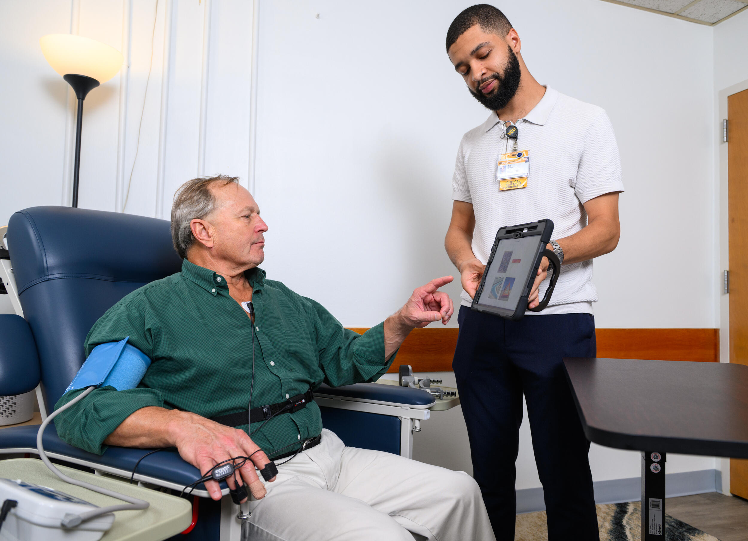 A photo of a man standing and a man sitting. The man standing is showing the sitting man an iPad with information on it.
