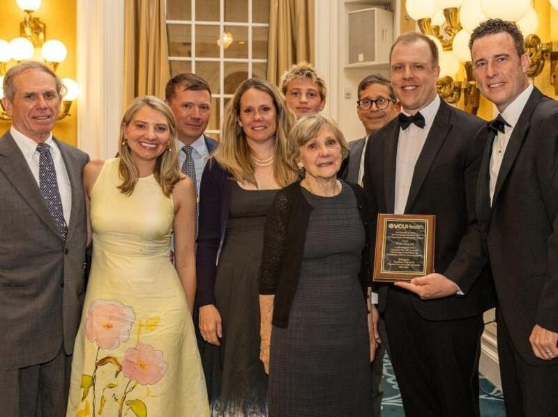A group photo of nine people. The second man to the right is holding an award. 