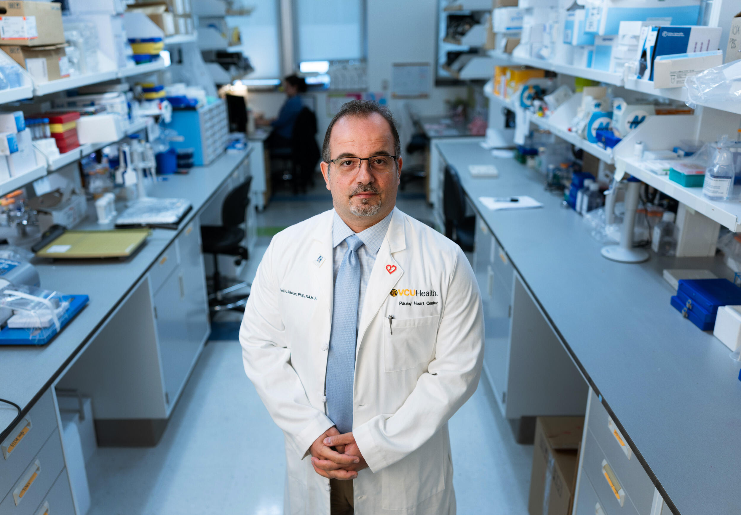A man in a white lab coat stands in a lab environment.