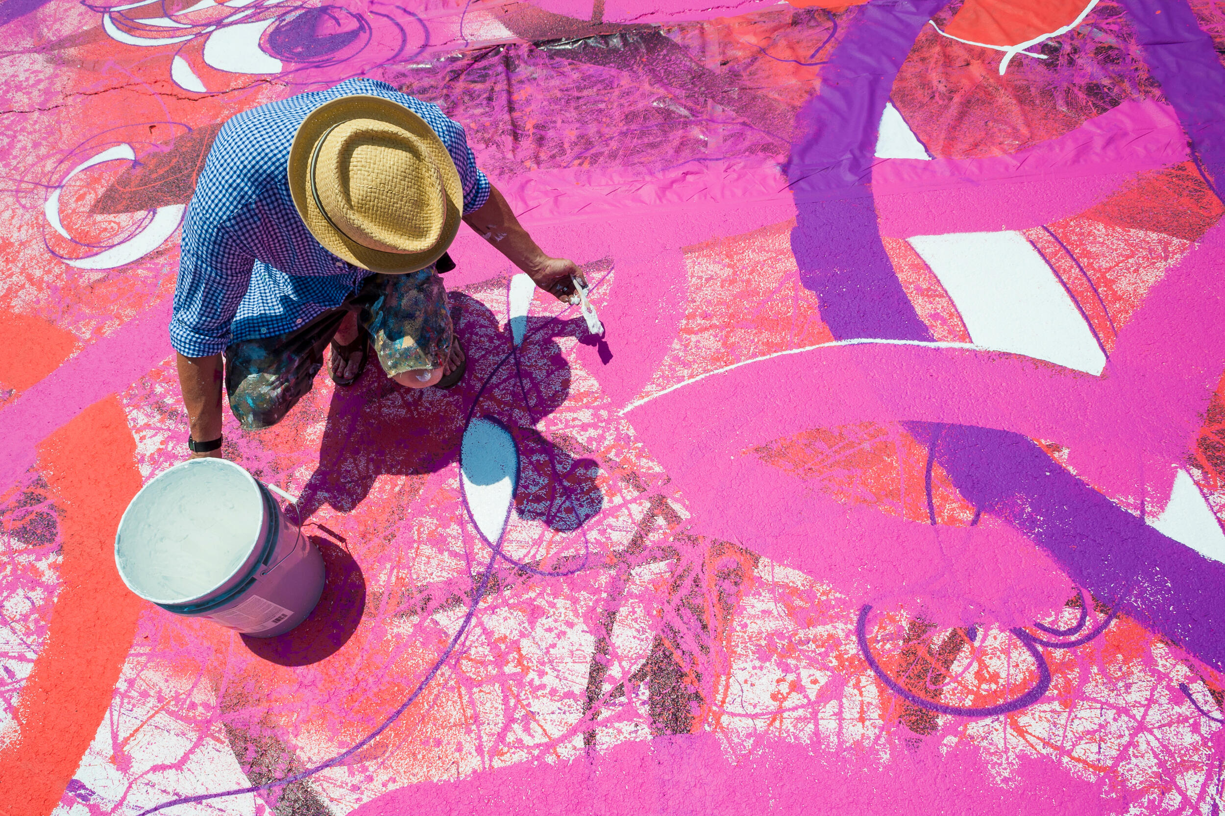 A man in a hat and coveralls with a paintbrush paints a colorful mural underfoot on pavement outside a building