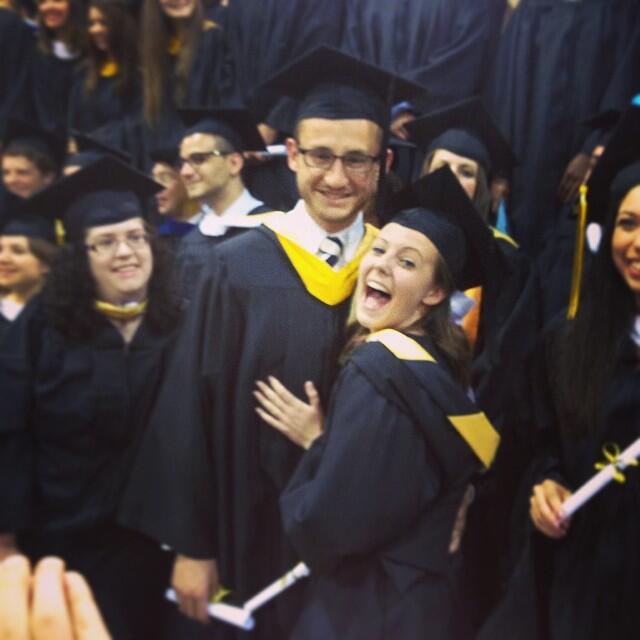 A photo of a man and woman wearing graduation cap and gowns standing in a crowd with other graduates. The woman has her hand resting on the man's torso. Both the man and woman are smiling. 