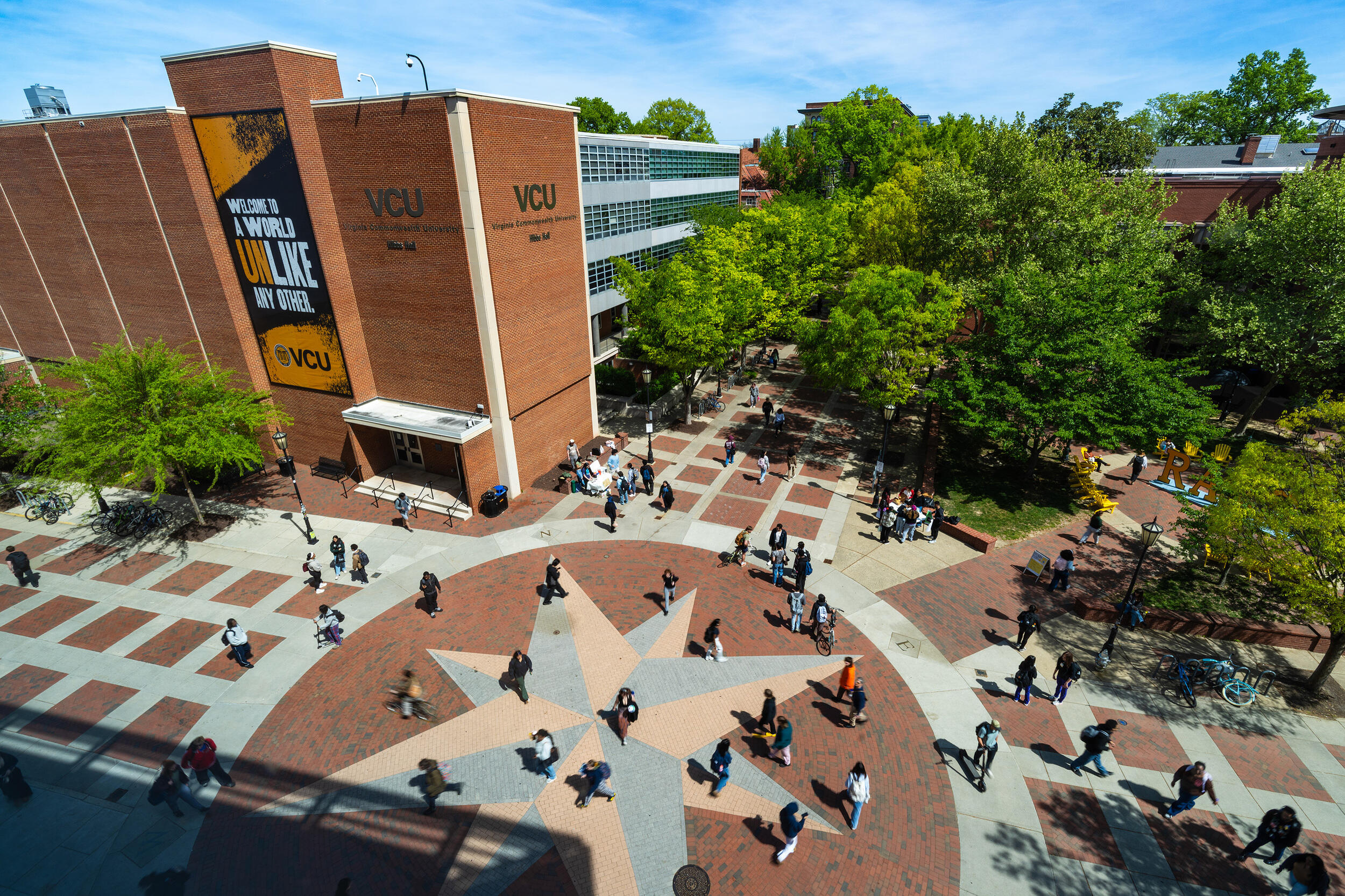 An aerial photo of the Comss plaza on VCU's Monroe Park campus. On the ground bricks in three different colors to look like the star in the middle of a compass. There are people walking across the plaza and the trees all have green leaves. 