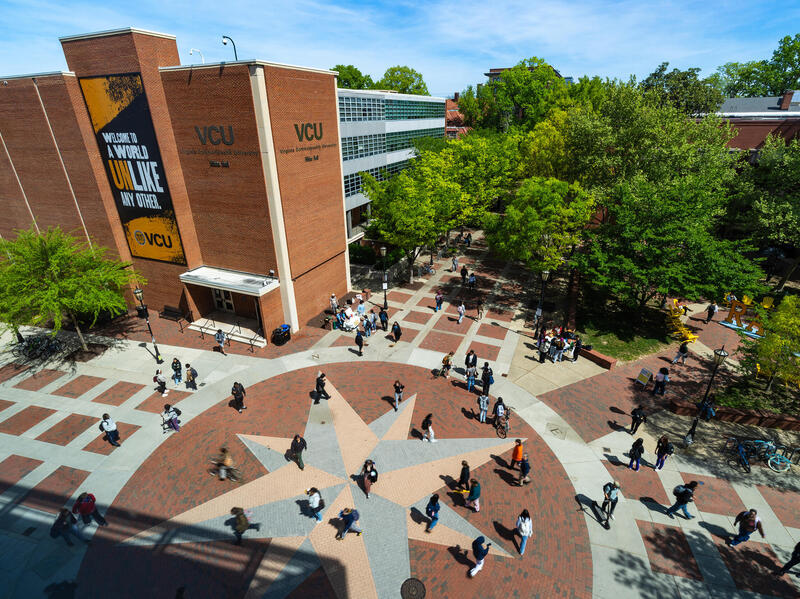 An aerial photo of the Comss plaza on VCU's Monroe Park campus. On the ground bricks in three different colors to look like the star in the middle of a compass. There are people walking across the plaza and the trees all have green leaves. 