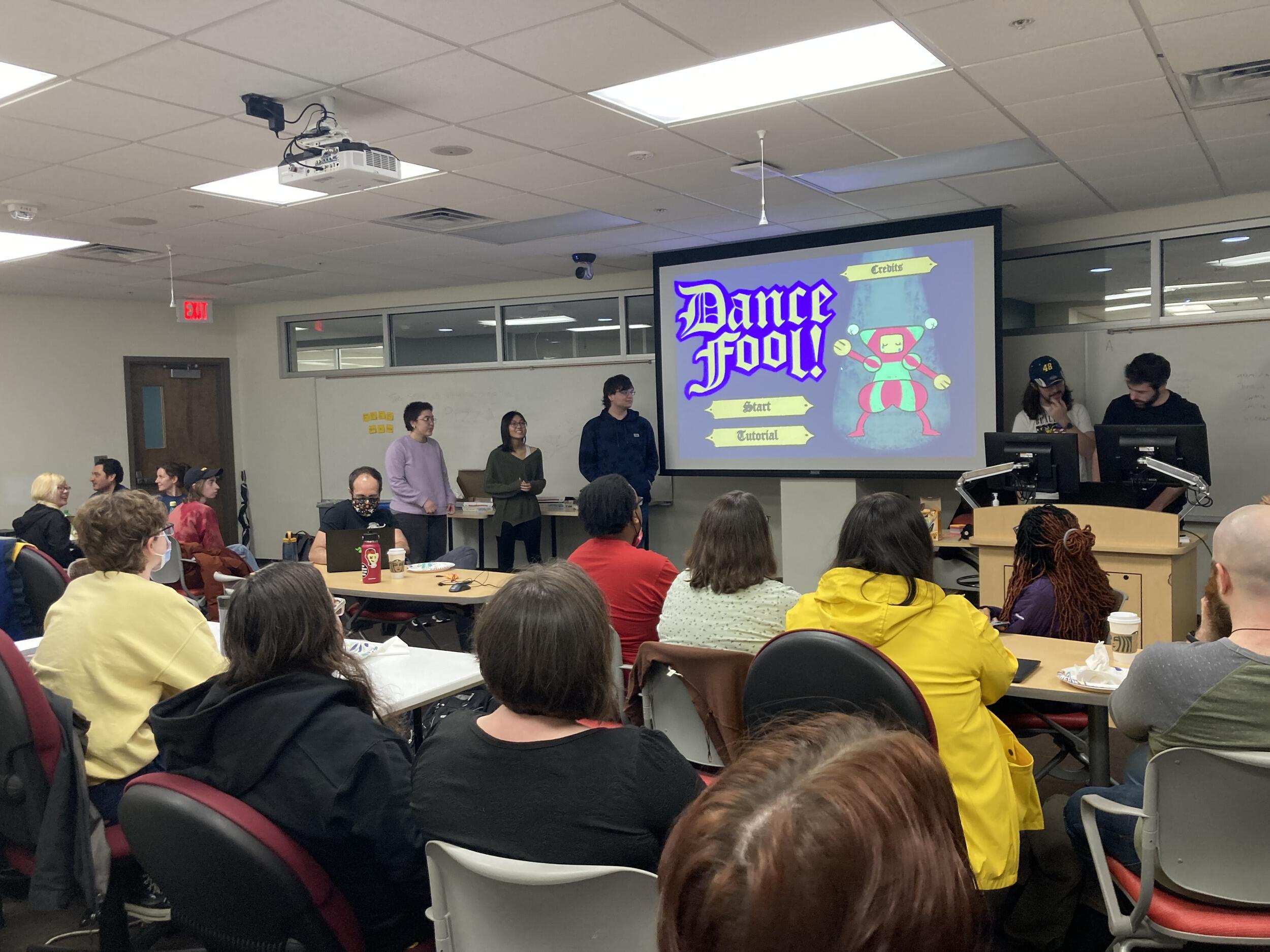 A photo of five people standing at the front of a classroom with a projector screen showing the image of a the start screen for a video game. The room is full of people sitting at tables watching the group presenting. 