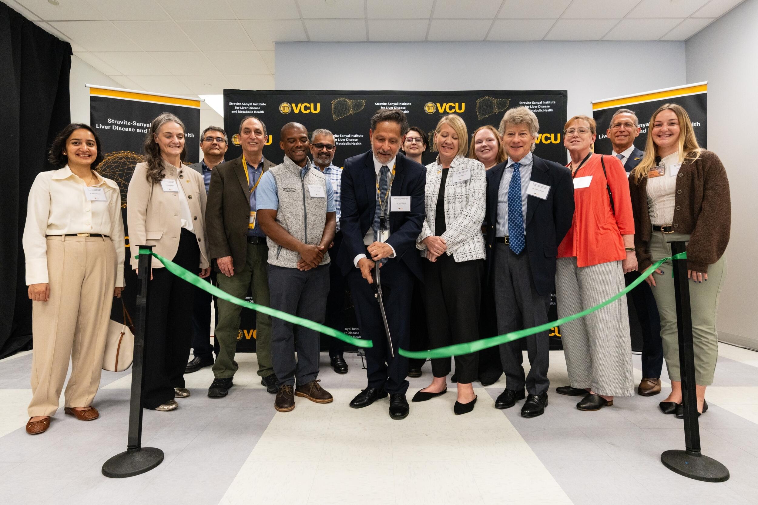 A group photo of 14 people standing behind a green ribbon. A man in the center with giant scissors is cutting the ribbon.
