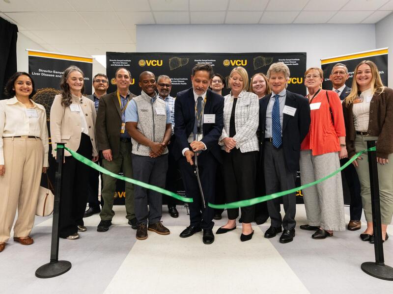 A group photo of 14 people standing behind a green ribbon. A man in the center with giant scissors is cutting the ribbon.