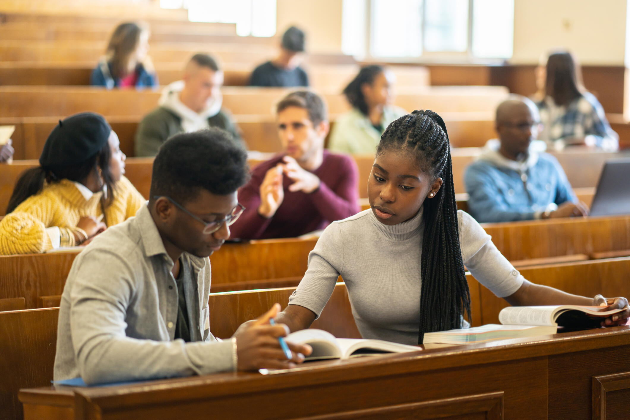 A photo of a row of lecture seats in a college classroom. There are students scattered throughout the rows. In the front is a Black man and woman who are reading off of the same book. The woman is speaking and the man is holding a pencil and one page of the book. 