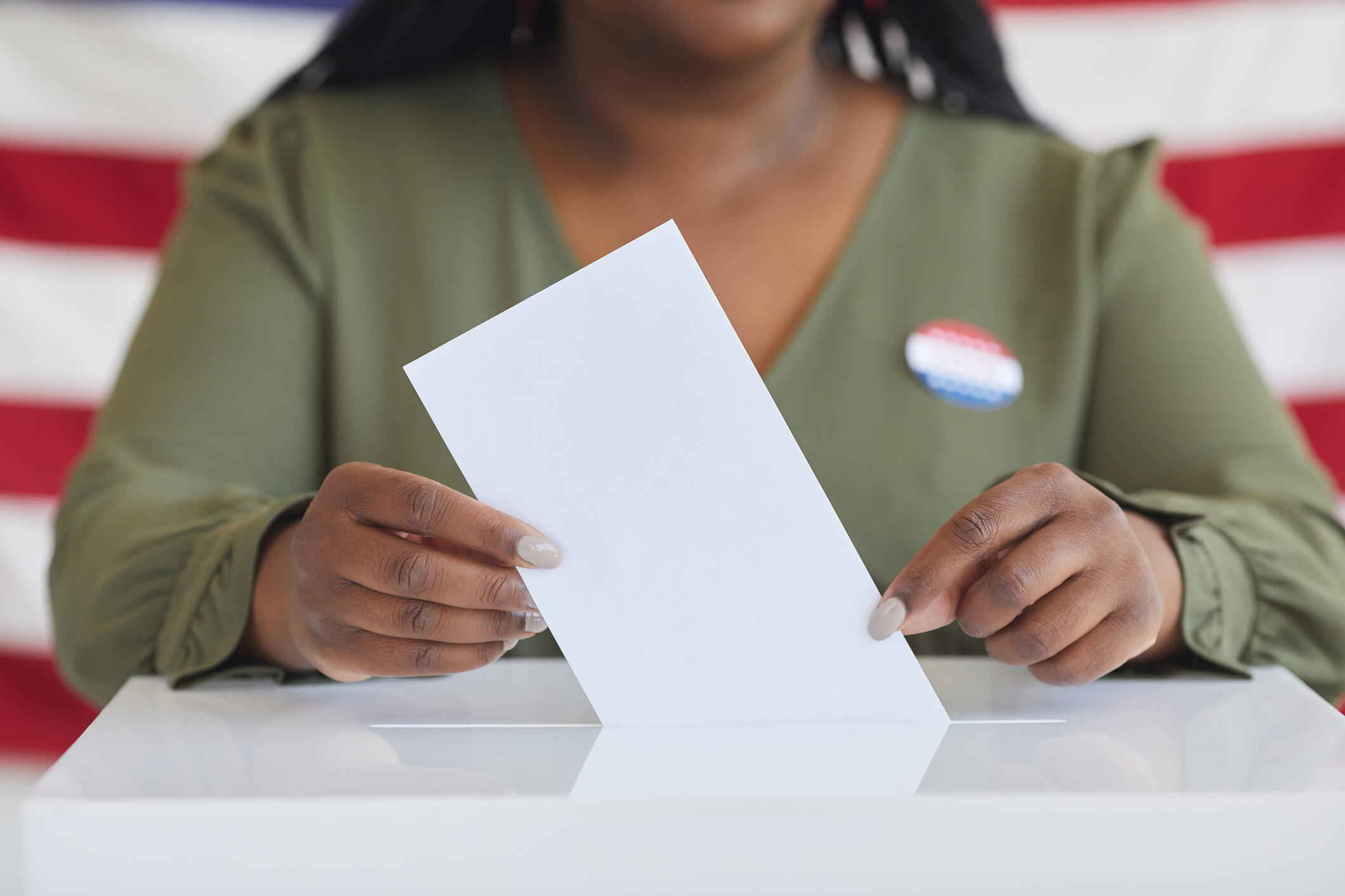 A photo of a woman's torso putting a piece of paper in a ballot box. 