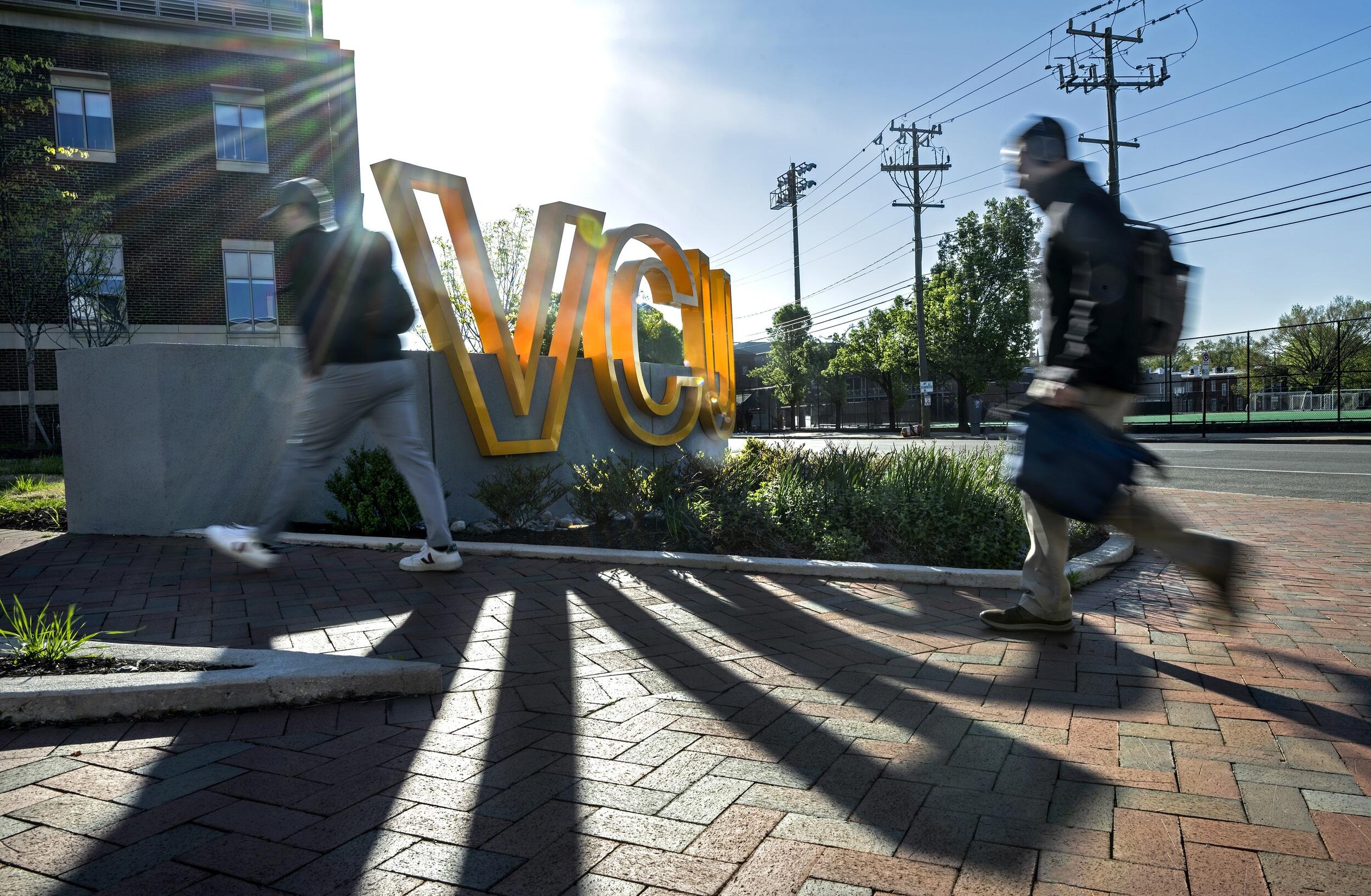 A photo of two people walking past a sign made of giant letters that spell out \"VCU\" 