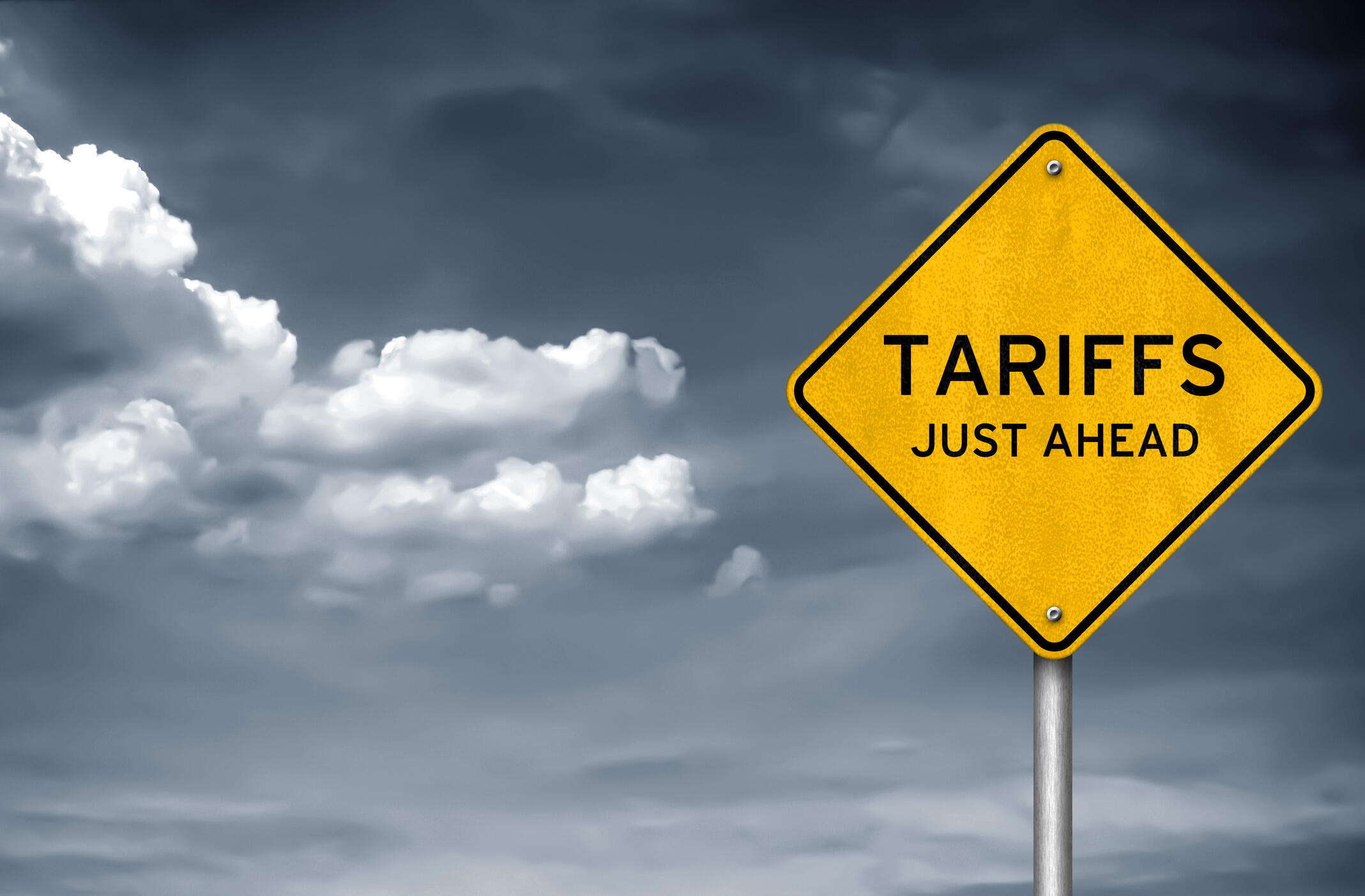 A photo of a yellow diamond shaped traffic sign that says \"TARIFFS JUST AHEAD\" in black letters. 