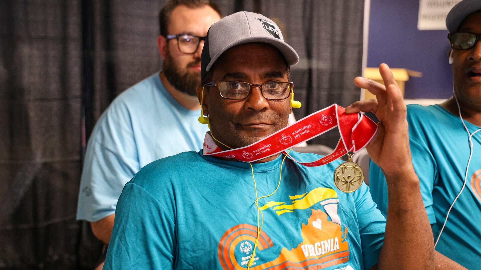 A photo of a man wearing glasses, a blue t-shirt, and a gray baseball hat holding up a gold medal that is around his neck. The medal is hanging off of a red ribbon. Next to him is a man wearing the same shirt and hat looking at the man wearing the medal, and behind the two men is another man wearing a light blue t-shirt and glasses. 
