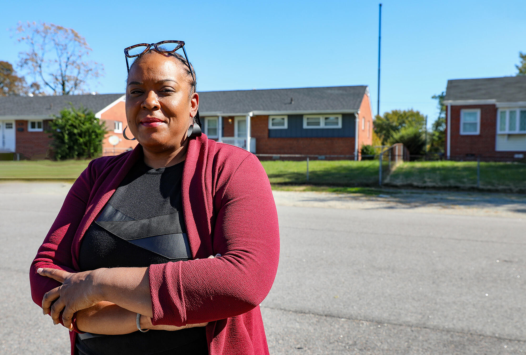 A photo of a woman from the waist up. Her arms are crossed against her chest. She is standing in the street in front of three houses. 