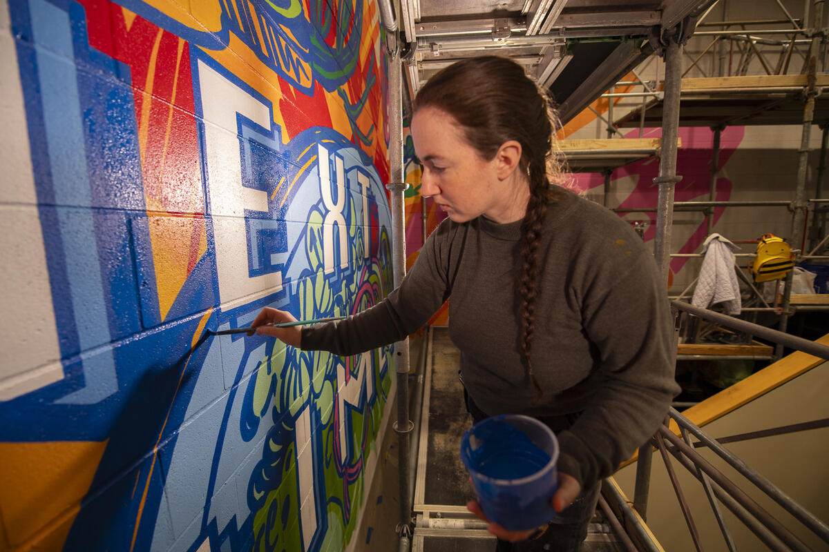 A photo of a woman holding a container of blue paint and painting on a wall. 