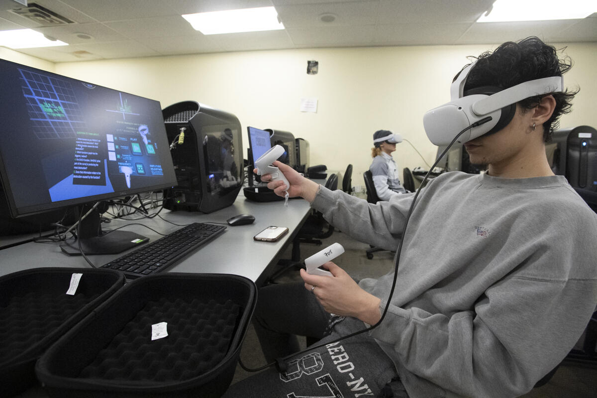 A photo of a man wearing a VR headset and holding two remotes while sitting in front of a computer screen. 