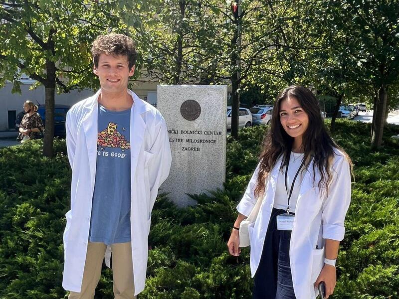 A photo of a man and woman in lab coats standing outside in front of some bushes and trees. 