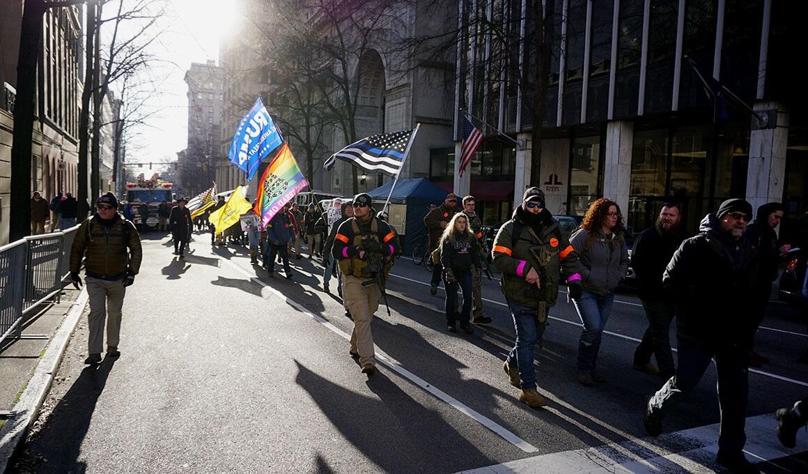 Group of people walking down the street, some holding flags or guns. 