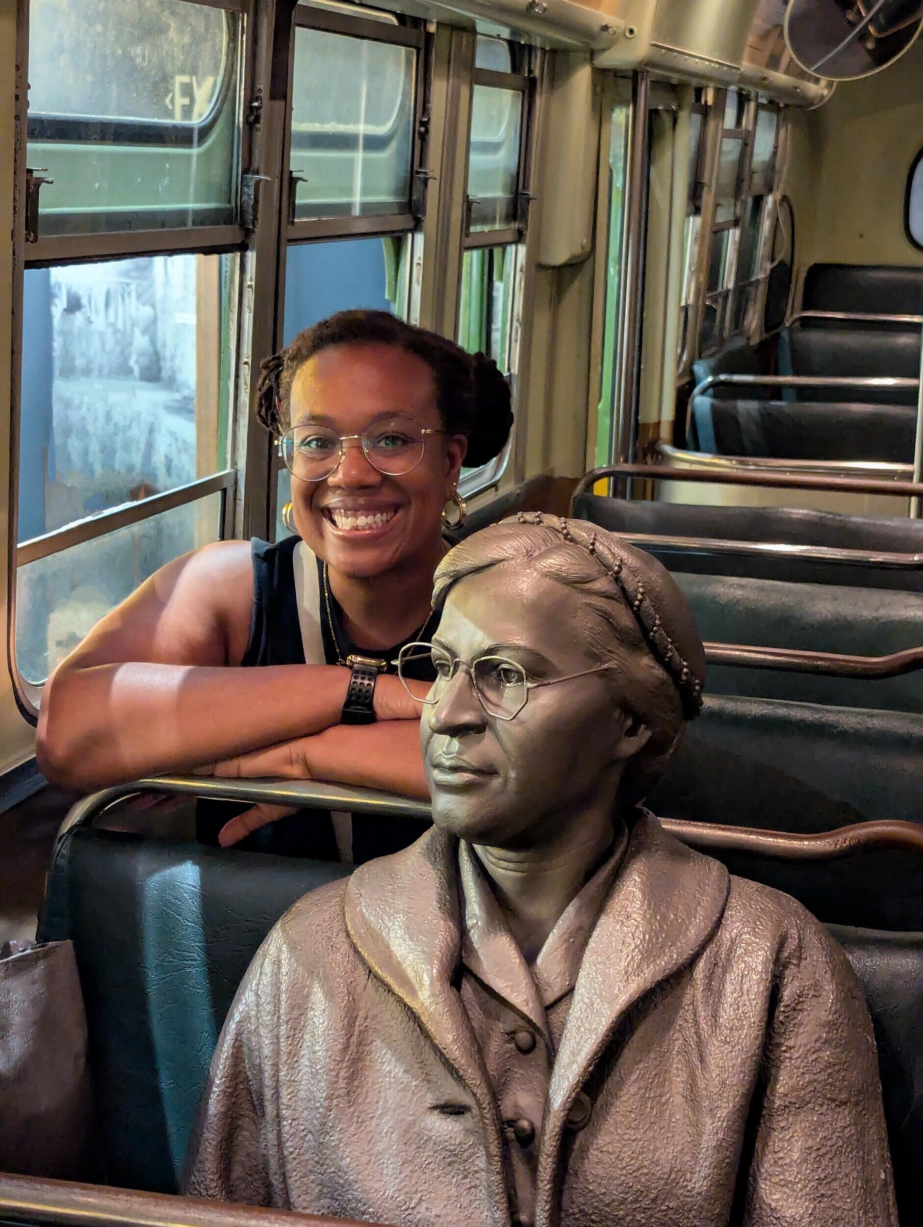 A photo of a woman sitting in a bus seat behind a statue of Rosa Parks.
