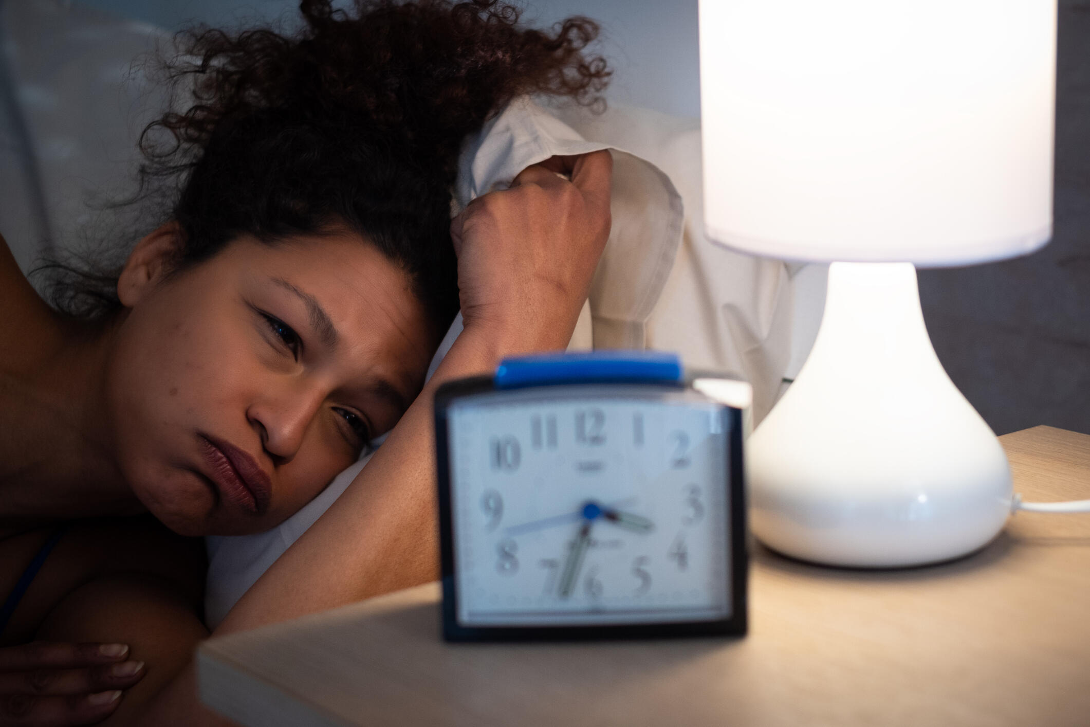 A woman laying in bed with a frustrated expression on her face as she stares at a clock next to her on a nightstand. 