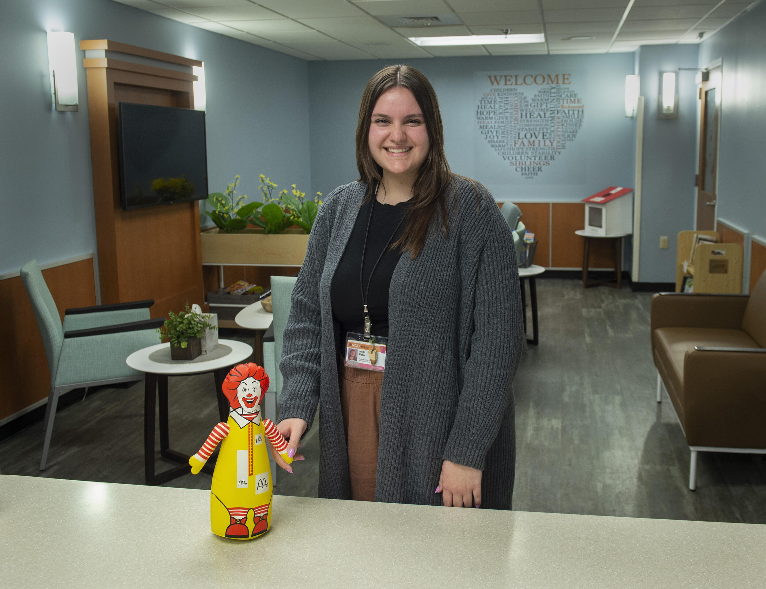 A woman standing in a room smiling. In front of her is a table with a red and yellow clown toy. 