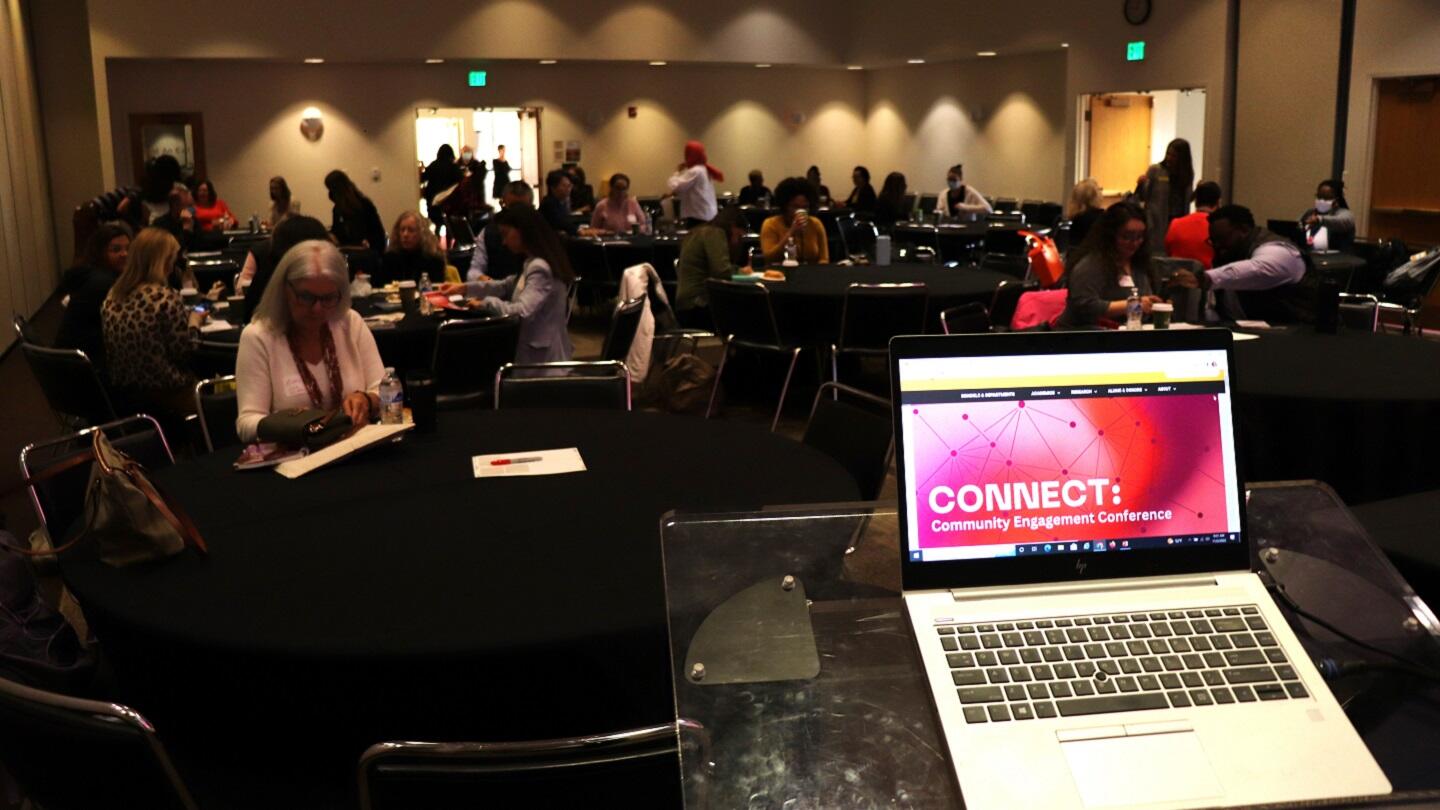 A laptop that says \"CONNECT: Community Engagement Conference\" on the screen. It is sitting on a podium in front of a room full of people. 