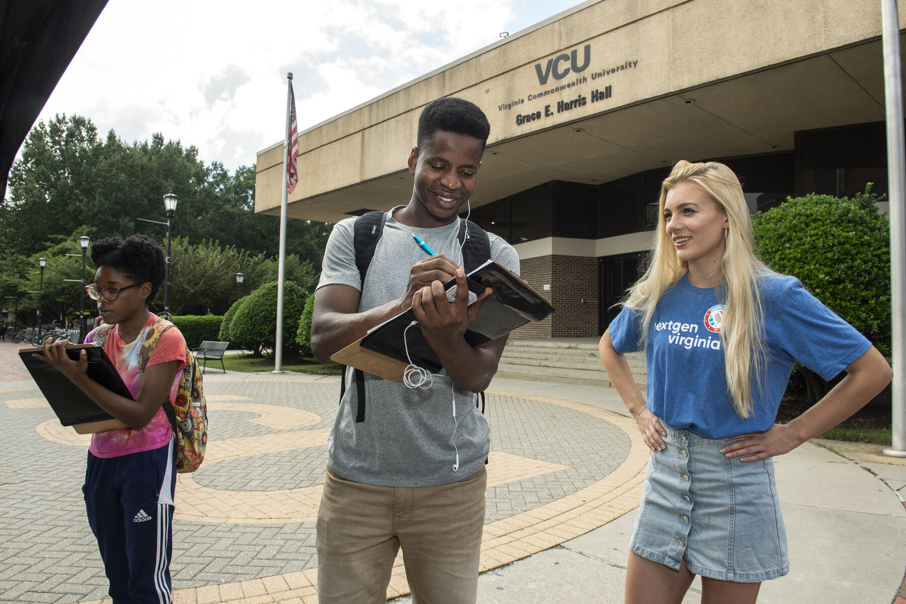 A woman in a pink shirt and a man in a grey shirt sign papers on clipboards next to Grace E. Harris Hall, while a woman in blue T-shirt with "nextgen Virginia" written on it watches.