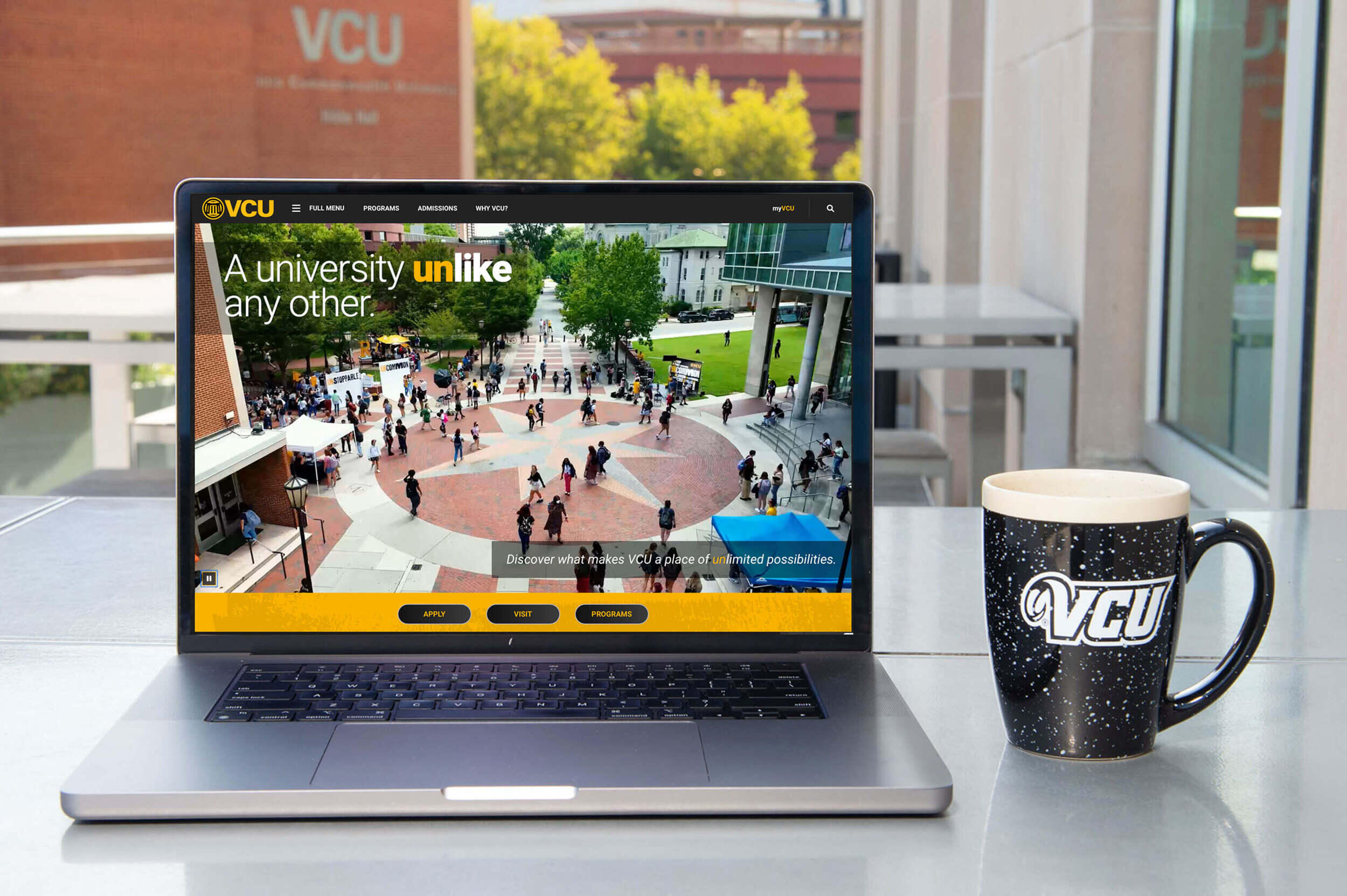 A laptop sitting on a table in front of a window. Next to the laptop is a coffee mug that says \"VCU\" in white letters. 