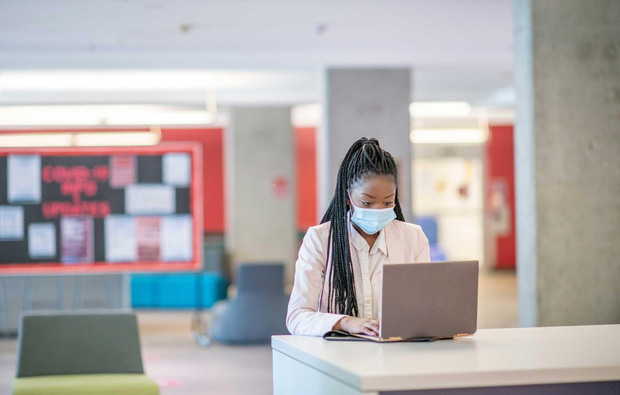 young black female college student sitting alone in a study space, wearing a face mask