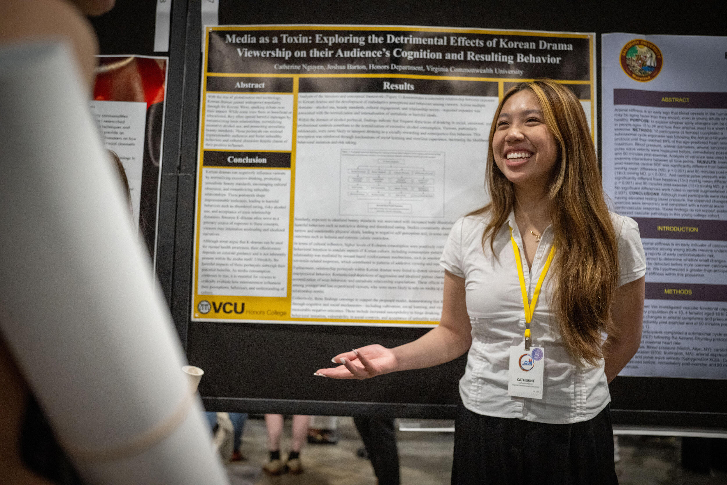 A photo of a woman standing in front of a research poster smiling and gesturing her left hand.