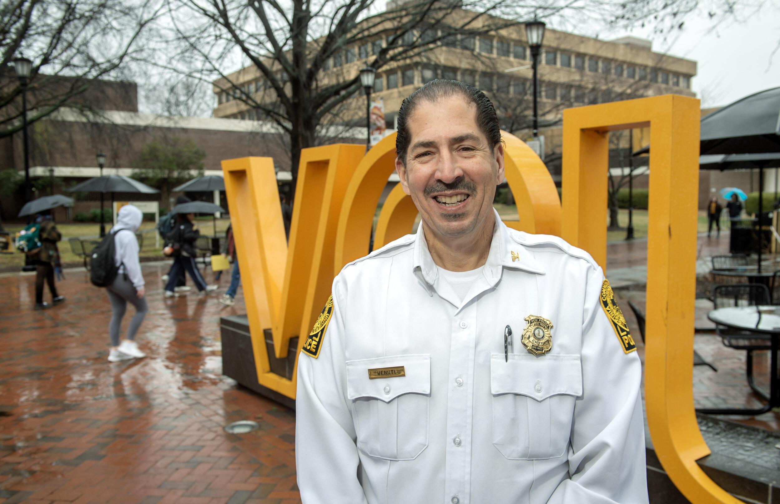 A photo of a man from the chest up. He is wearing a white police uniform shirt and a gold police badge. Behind him is a yellow sign that says \"VCU\" in large yellow letters. 