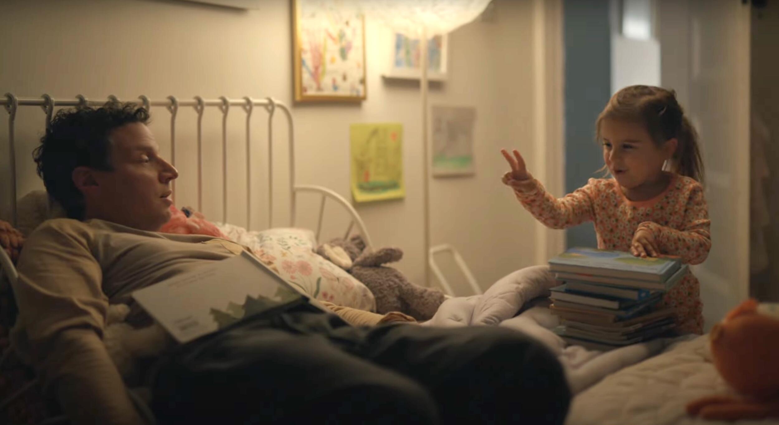 A photo of man laying on a bed. Standing next to the bed is a little girl holding up one hand with two fingers outstretched. 