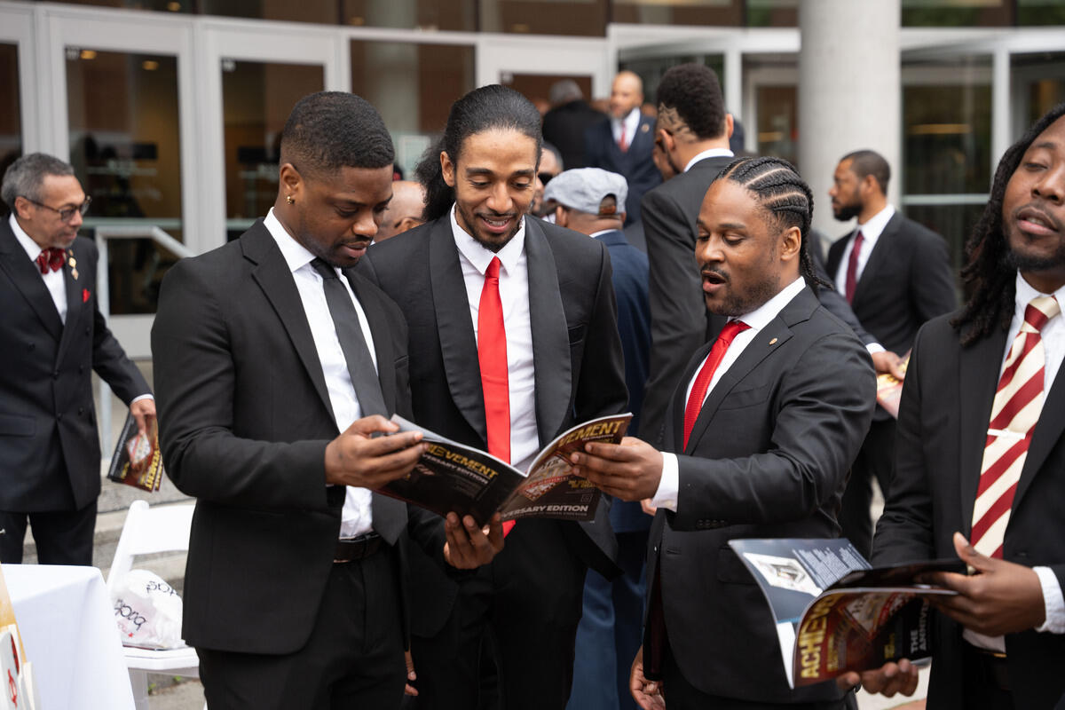 A photo of three men looking at a magazine together. Another man is holding a magazine next to them. A crowd of people is mingling behind the group of four men. 