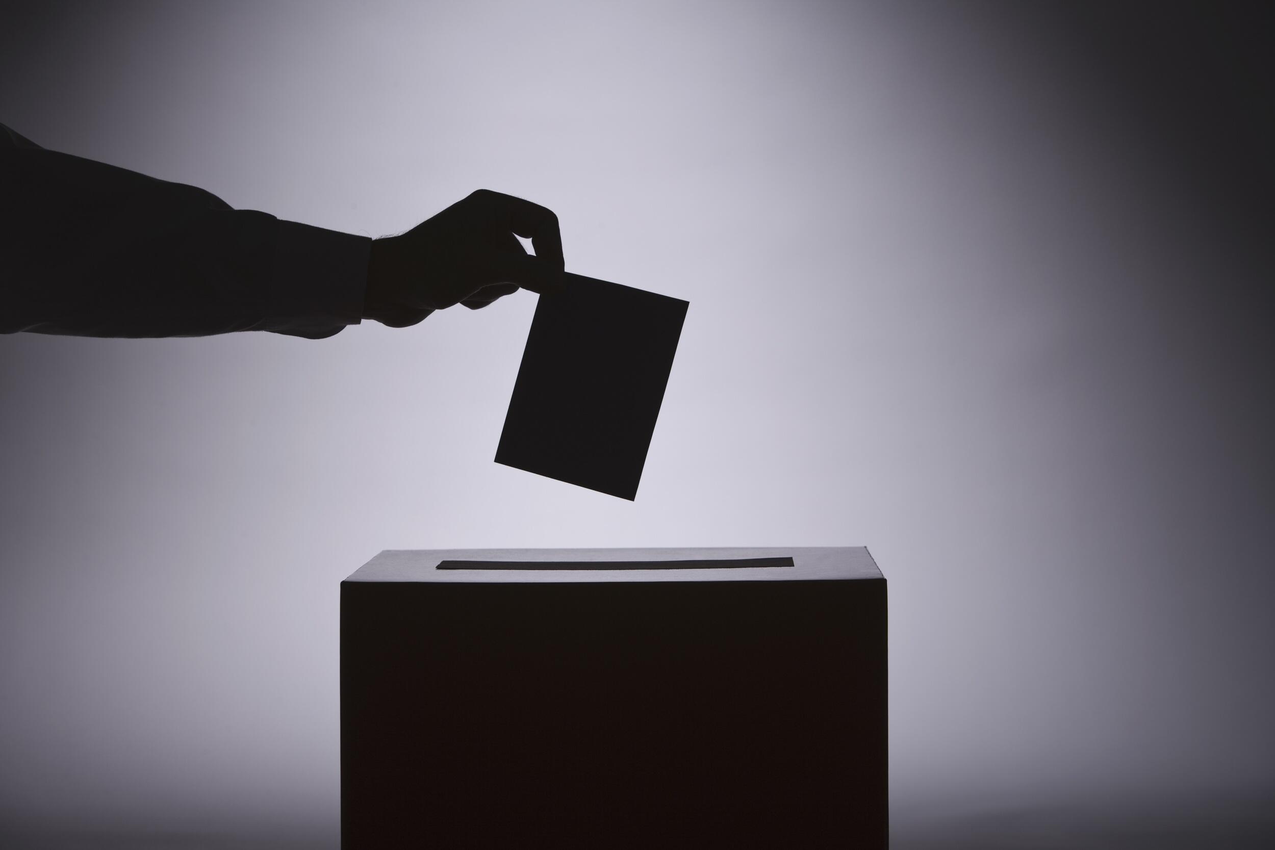 A hand holds a ballot over a ballot box.