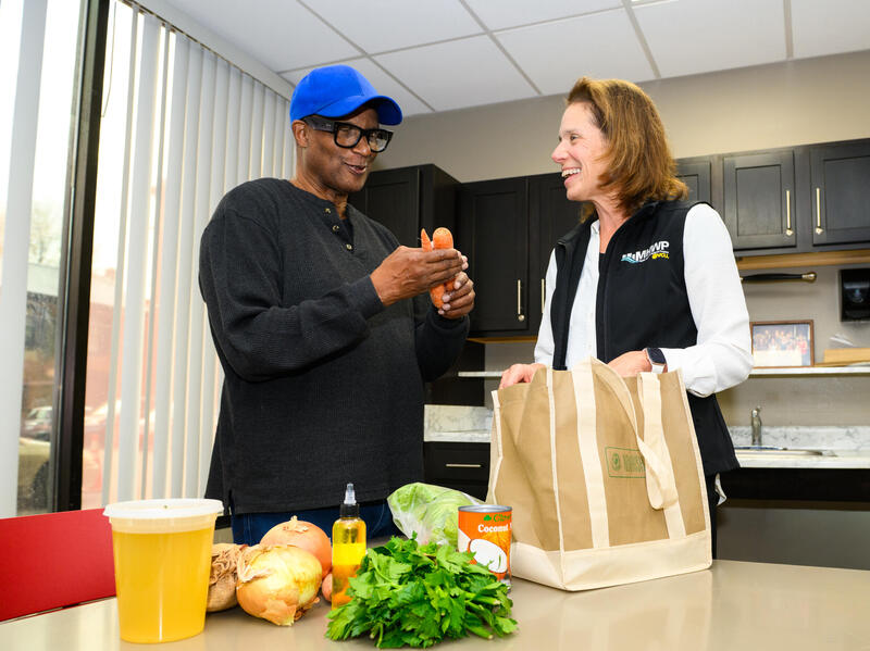 A photo fo a man and woman standing behind a table. The table has a reusable bag, produce, and a container of broth on it.